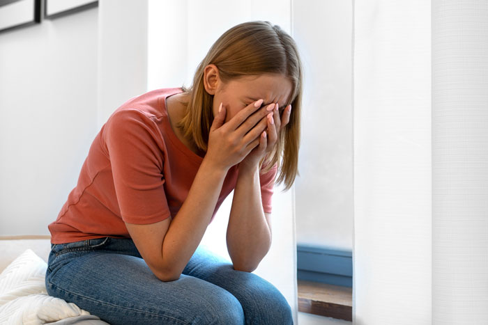 Young woman sitting indoors covering her face in distress after uncovering a boyfriend's wedding lie and secret life.