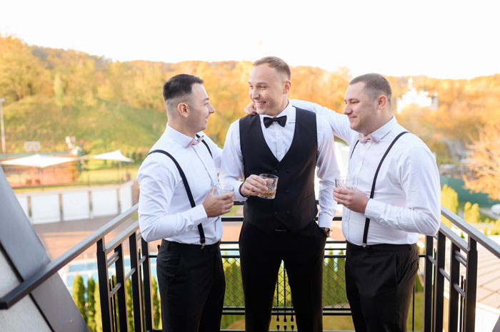 Three men in formal attire with suspenders and bow ties, socializing with drinks at an outdoor wedding event.