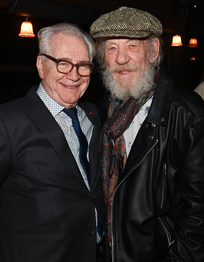 Brian C*x wearing glasses and a suit posing with a bearded man in a leather jacket and patterned scarf at an event.