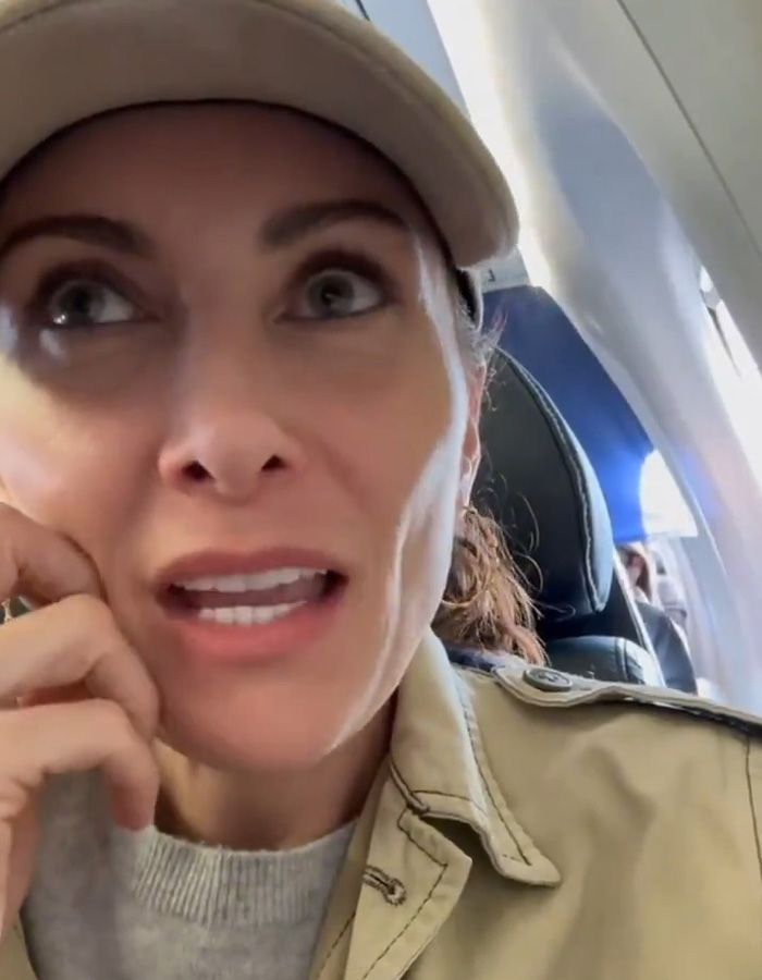 Broadway star speaking on a plane, wearing a cap and beige jacket, reacting to not being recognized by passengers.