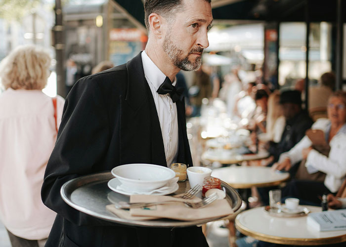 Man working as a server in a busy restaurant carrying a tray with dishes, focused on his job in a crowded dining area. Man working as a server in a busy restaurant carrying a tray with dishes, focused on his job in a crowded dining area.