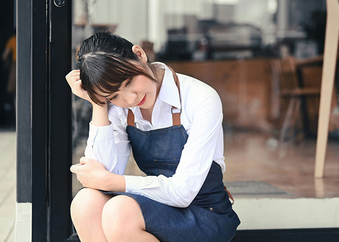 Young female server in a denim apron sitting outside a cafe looking contemplative about friendship and work value. Young female server in a denim apron sitting outside a cafe looking contemplative about friendship and work value.