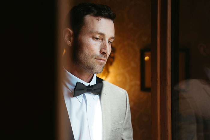 A groom in a light suit and black bow tie, looking contemplatively to the side, perhaps contemplating his wedding day decisions.