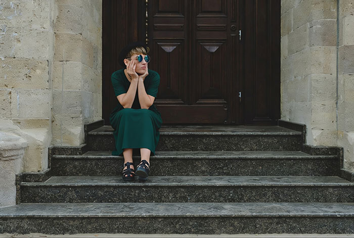 A young woman with a bob, sunglasses, and a black hat, wearing a green dress, sits on stone steps. She looks contemplative, possibly pondering a wedding dress choice.