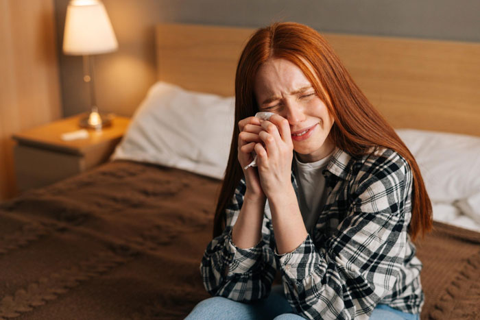 A young red-haired DIL crying with a tissue, upset in her bedroom, feeling sensitive and sad.