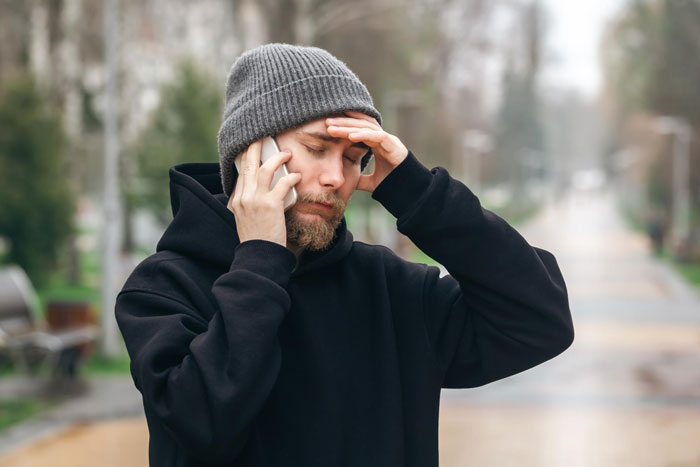 Man in black hoodie and gray beanie talking on phone outside, appearing frustrated after revenge on honking driver.
