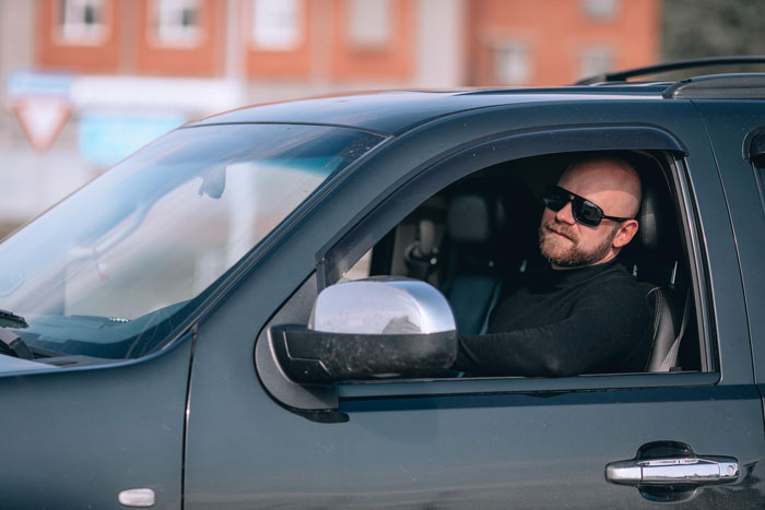 Man sitting in car wearing sunglasses showing a smirk after getting revenge on honking driver with ice-filled bin.