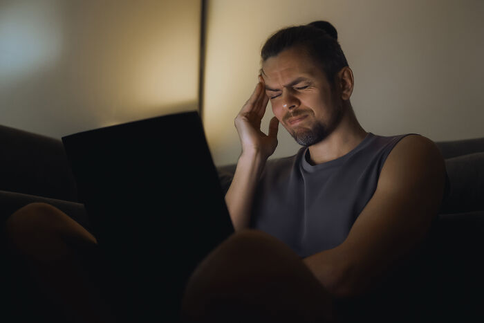 A man with a headache, eyes closed, sitting in a dim room with a laptop. He's accidentally finding out he was being cheated on.
