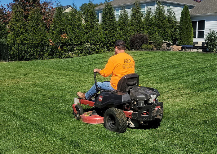 Man wearing orange shirt mowing green lawn on riding mower in a suburban backyard with tall trees and houses nearby
