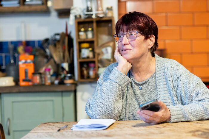 A pensive woman with glasses in a gray cardigan, holding a phone, sitting at a table. Relatable MIL stories.