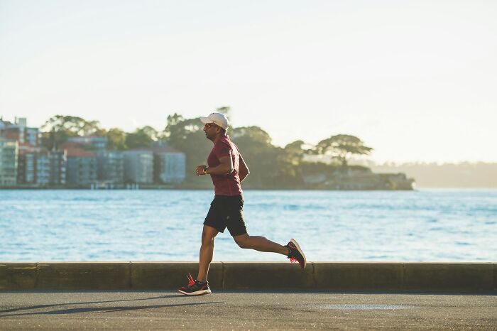 Man in a white cap, red shirt, and black shorts running along a waterfront. Reflects the cost of fitness.