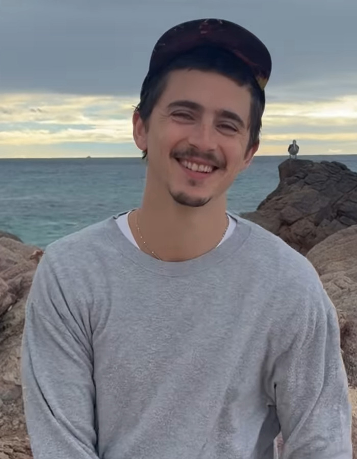 Young man smiling by the rocky shore with ocean background, related to internet divided over Charlize Theron ballet comment.
