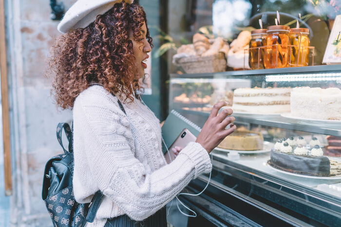 A young woman with curly hair and a beret gazes excitedly at cakes in a bakery window, embodying surviving adult life.
