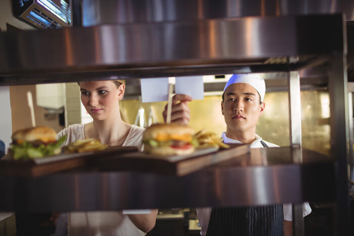 Two restaurant staff preparing and delivering orders with silent satisfaction in a busy kitchen setting around burgers and fries.