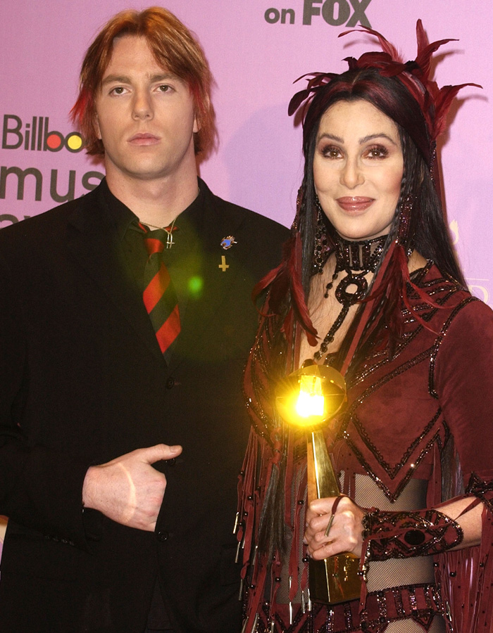 Cher sorrindo em uma roupa vermelha detalhada segurando um prêmio ao lado de um homem de terno preto e gravata listrada em um evento musical.