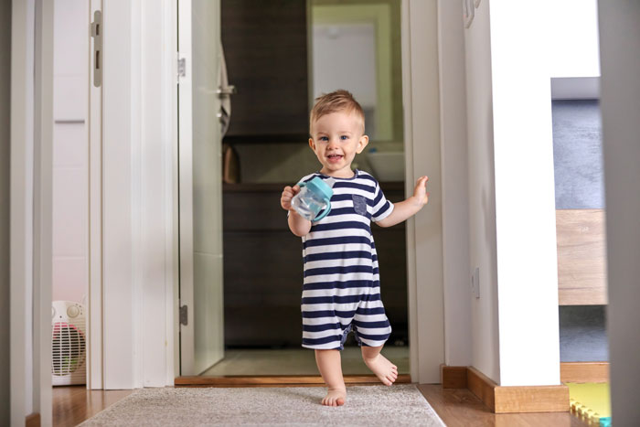 3-year-old toddler in striped pajamas walking inside a home holding a sippy cup, escaping from parent supervision.