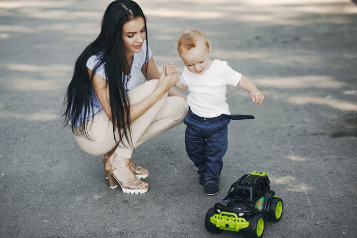 Woman helps 3YO toddler walk outside near toy truck, illustrating a young child escaping home to neighbor&rsquo;s house scenario.