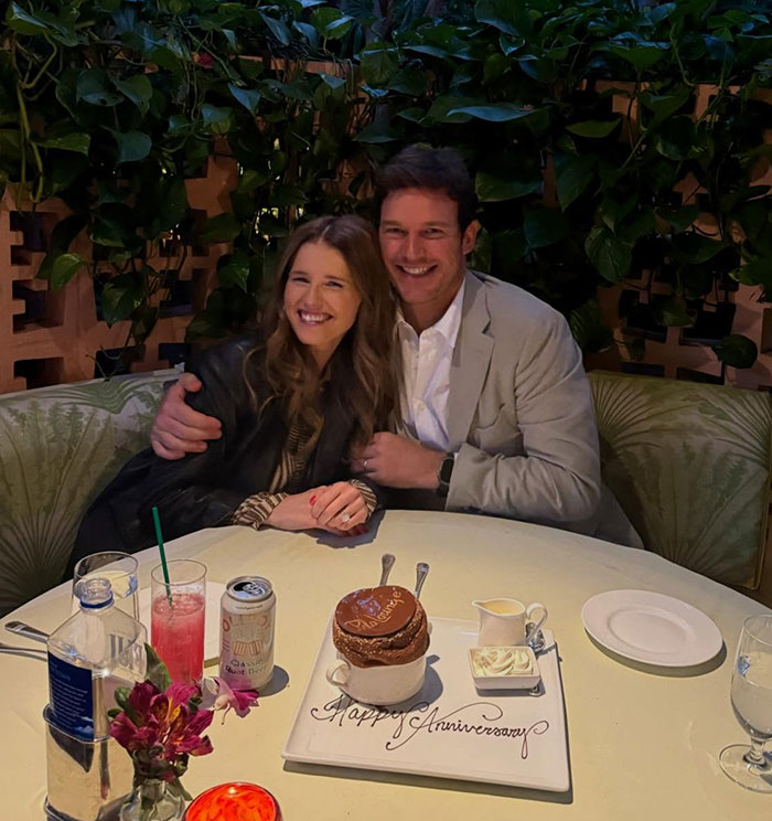 Katherine Schwarzenegger and husband Chris Pratt smiling at a dinner table with anniversary dessert and drinks.