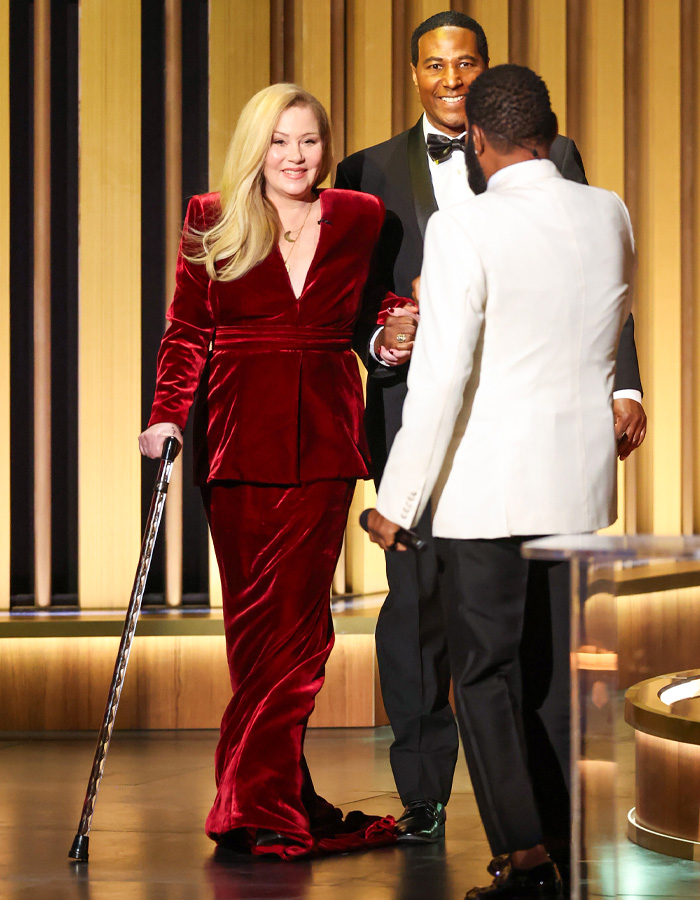 Christina Applegate in a red velvet dress using a cane, meeting others during a formal event amid hospitalization concerns.