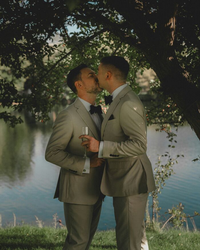 Two grooms in beige suits sharing a kiss under a tree by a lake, capturing a moment in wedding stories.