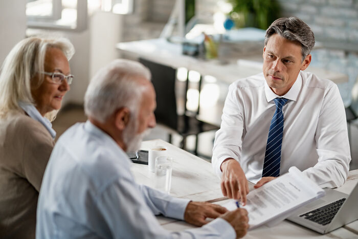 A male lawyer in a white shirt and striped tie, reviewing documents with an older couple, hinting at family crimes.