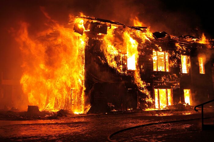 A large house engulfed in huge, bright orange flames and smoke at night, symbolizing unhinged crimes.