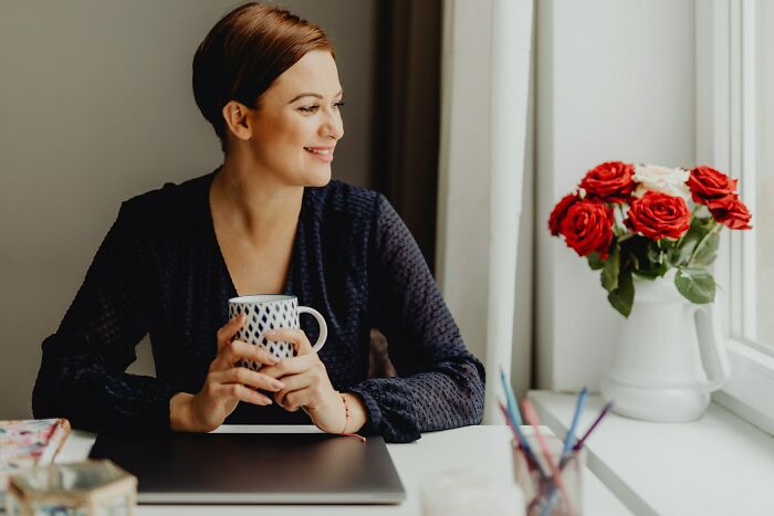 Woman holding a mug and smiling thoughtfully while sitting near a window with a vase of red roses, reflecting on bullies.