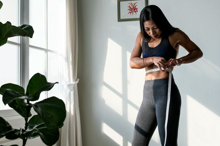 Woman measuring waist with tape indoors by window with plants, illustrating horrendous things committed by best friends concept.