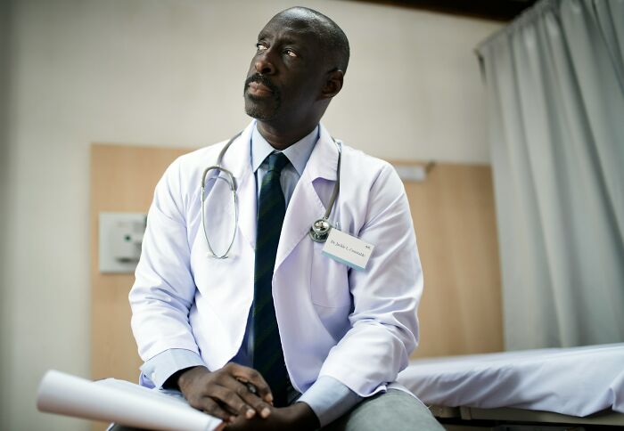 Black male gynecologist wearing white coat and stethoscope, sitting contemplatively in a clinical setting.