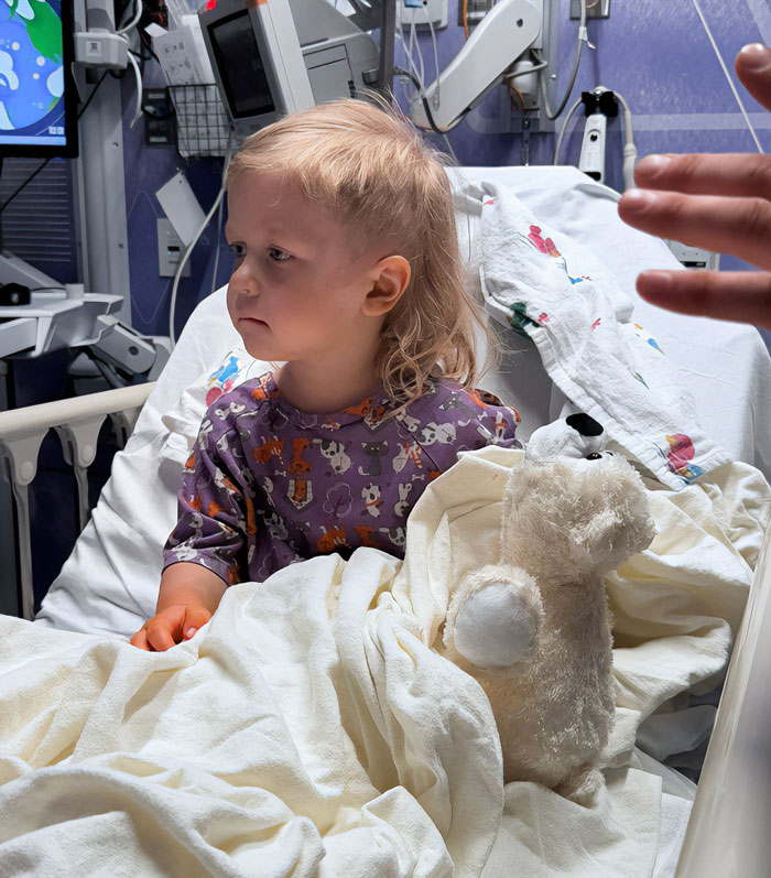 Toddler sitting in hospital bed with stuffed animal, receiving care after incident involving police intervention at Walmart.