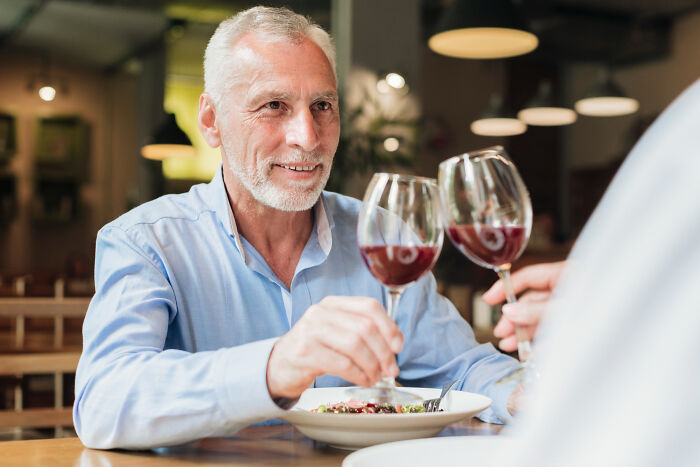 Older man in a blue shirt smiling and toasting red wine glasses during a mid-date at a cozy restaurant.
