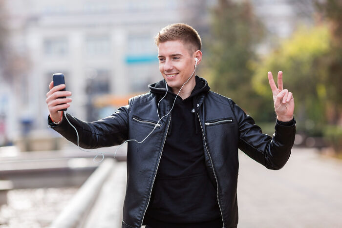Young man in a leather jacket taking a selfie outdoors, smiling and making a peace sign, capturing mid-date moments.