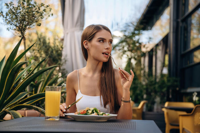 Young woman at an outdoor restaurant mid-date, thoughtfully eating salad and drinking orange juice, capturing unhinged reasons to leave.