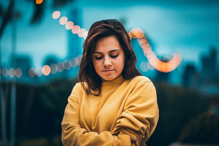 A young woman in a yellow sweatshirt with her eyes closed, deep in thought, against a blurred background of lights. Dads raising daughters.