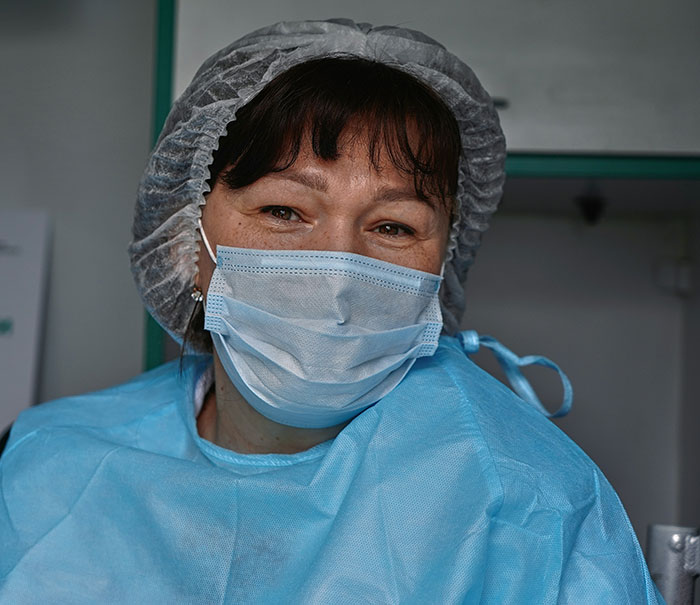 A female medical professional in scrubs, mask, and hairnet, smiling with her eyes. Sharing patient stories.