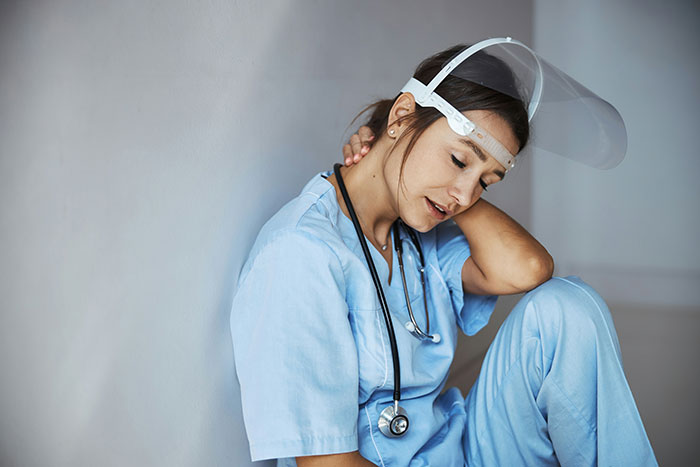 A tired medical professional in blue scrubs and a face shield, head down, rubbing her neck, exhausted from patient stories.