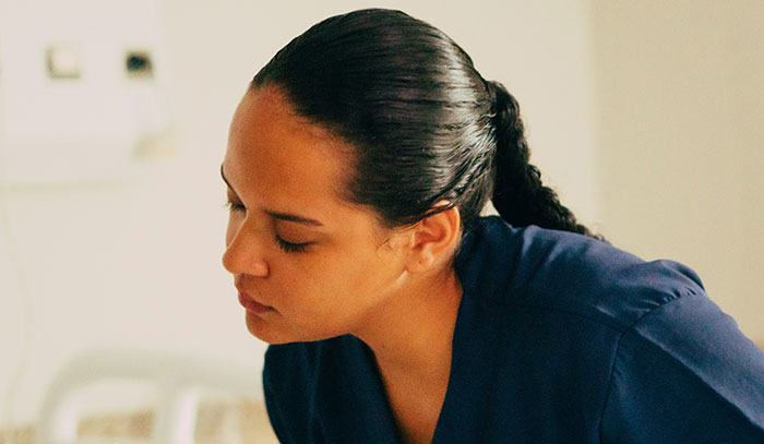 A medical professional, a woman with dark hair in a ponytail and a navy scrub top, looks down, sharing patient stories.