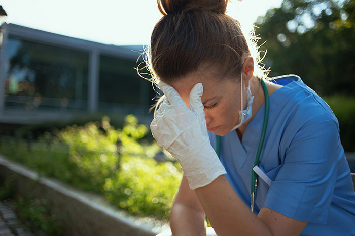 A tired medical professional in blue scrubs and white gloves, head in hand, reflecting on patient stories outside.