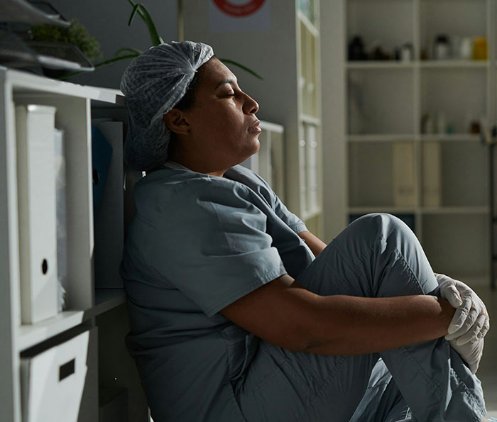 A medical professional, wearing scrubs and a hair net, sits slumped against a shelf, eyes closed, reflecting on patient stories.