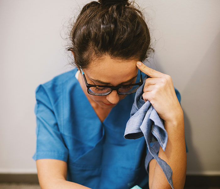 A medical professional in blue scrubs, appearing distressed, holds a cloth and touches her forehead. Patient stories can be intense.