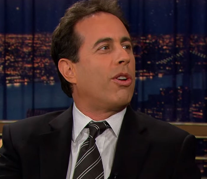 Comedian in a dark suit and striped tie speaking during a late-night show about disturbing celebrity facts.