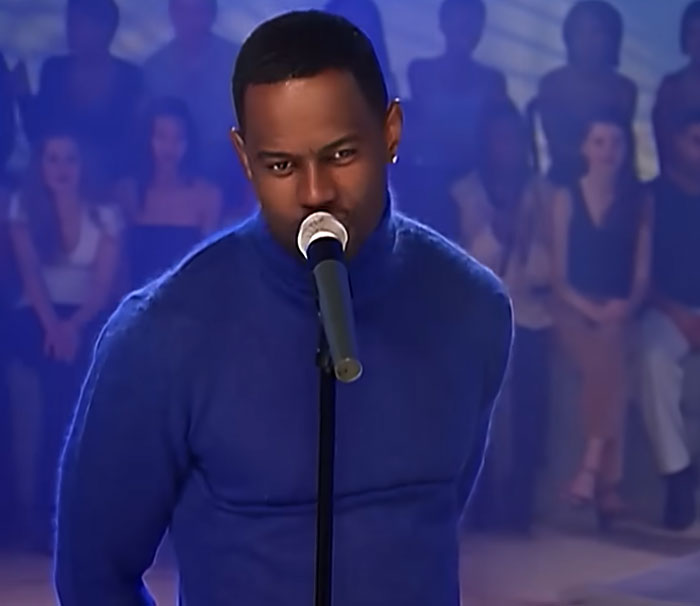 Man in a blue sweater speaking into a microphone with a seated audience in the background, related to disturbing celebrity facts.