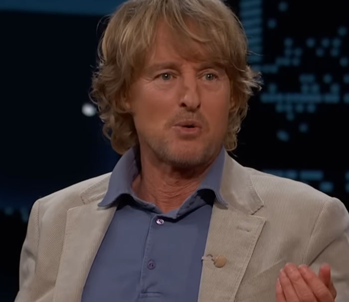 Actor with light brown hair wearing a beige jacket and blue shirt, speaking about disturbing celebrity facts on a dark set.