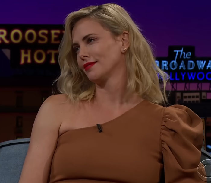 Blonde woman in a brown one-shoulder dress speaking on a late-night talk show about disturbing celebrity facts.