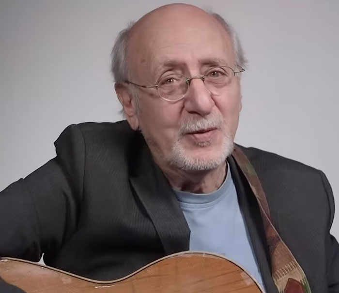 Elderly man wearing glasses and a blazer, playing guitar in a casual indoor setting with a neutral background.