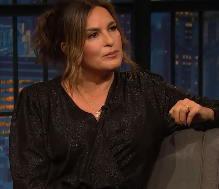 Woman in a black top seated on a couch, sharing disturbing celebrity facts in a dimly lit studio setting.