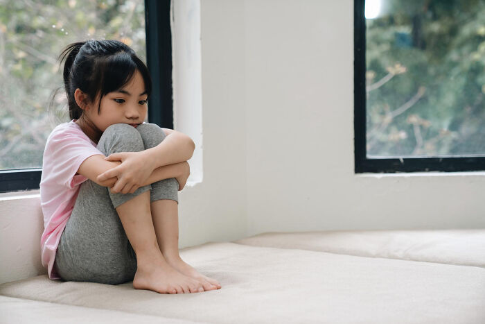 Young girl sitting alone by the window, looking sad and withdrawn, illustrating the impact of horrible things parents say.