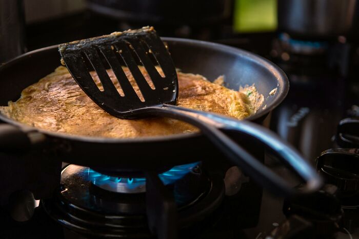 Spatula flipping a golden omelette in a black frying pan over a blue gas stove flame in a kitchen.