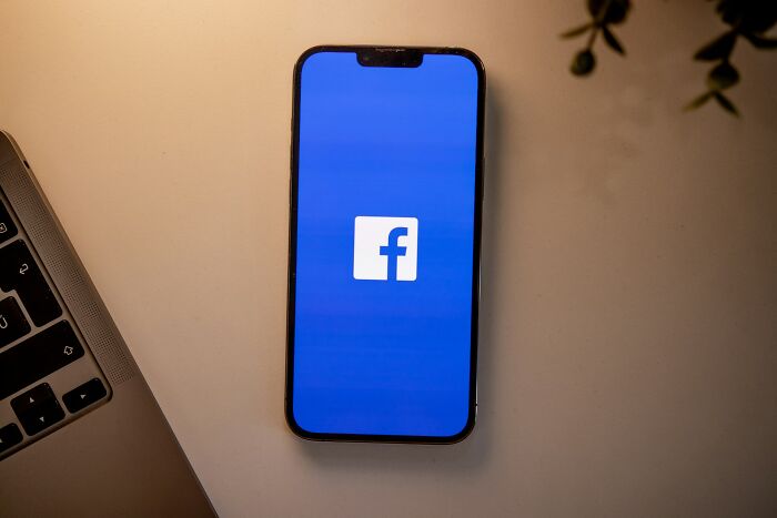 A smartphone displaying the Facebook logo on a blue screen, next to a laptop keyboard. Stories about MIL.