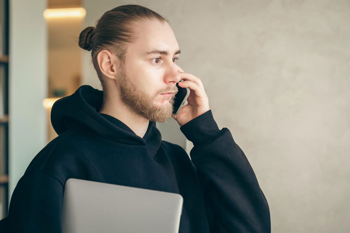 Young man with a beard in a black hoodie talking on phone while holding a laptop in a modern indoor setting